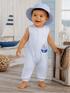 Smiling baby in a striped romper and blue hat stands on a wooden deck with nautical decor in the background.