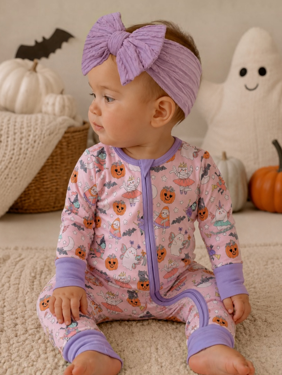 Baby in Halloween-themed pajamas with a purple bow, seated on a soft rug near pumpkins and a ghost decoration.