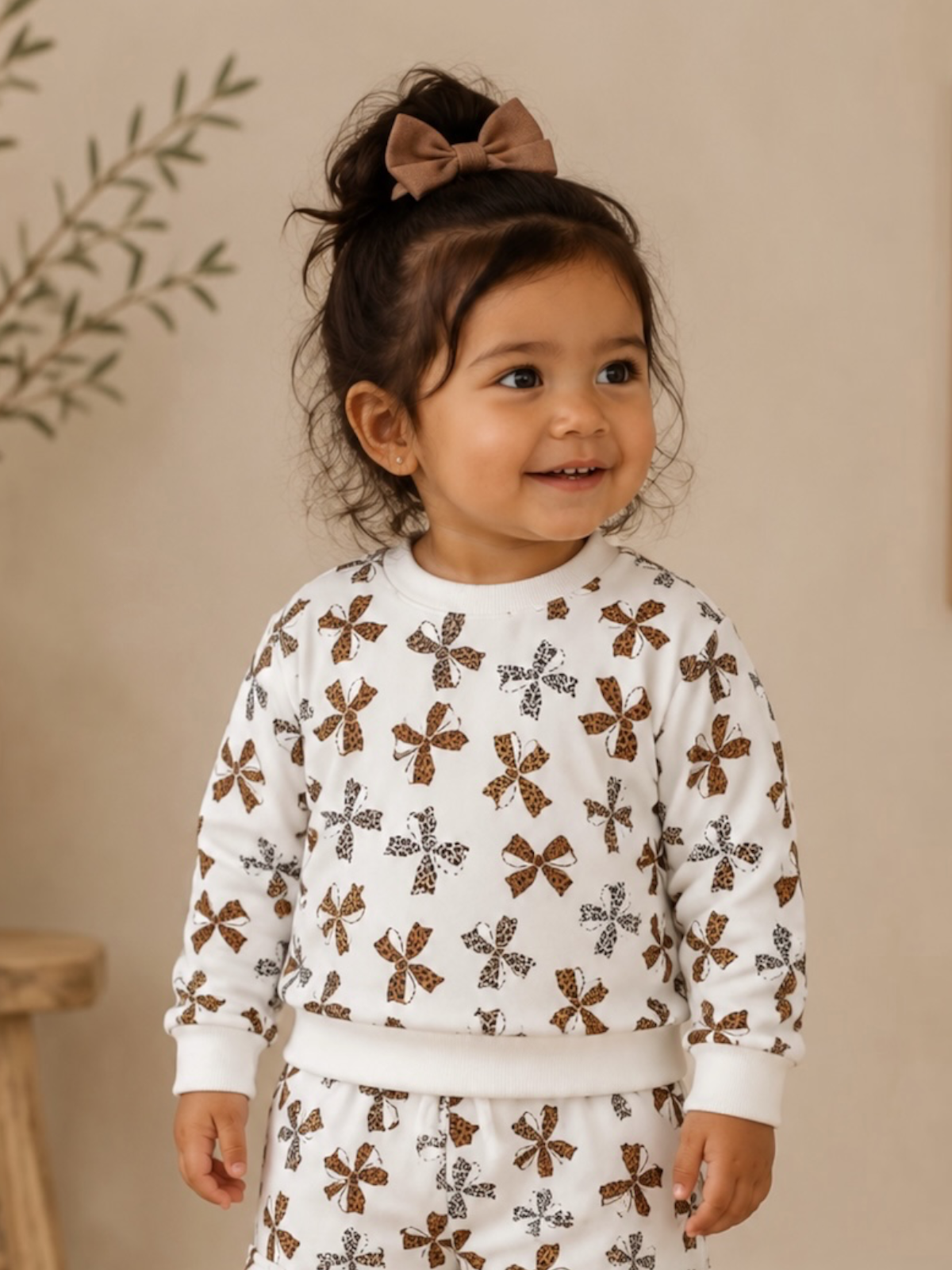 Smiling toddler in a patterned outfit with a bow in her hair, standing against a soft, neutral background.