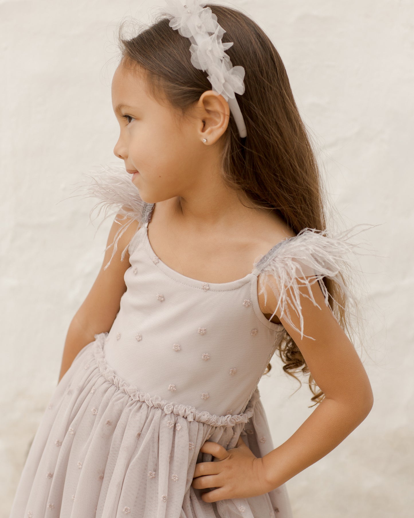 Young girl in a light dress with feather details, smiling and wearing a floral headband against a neutral background.
