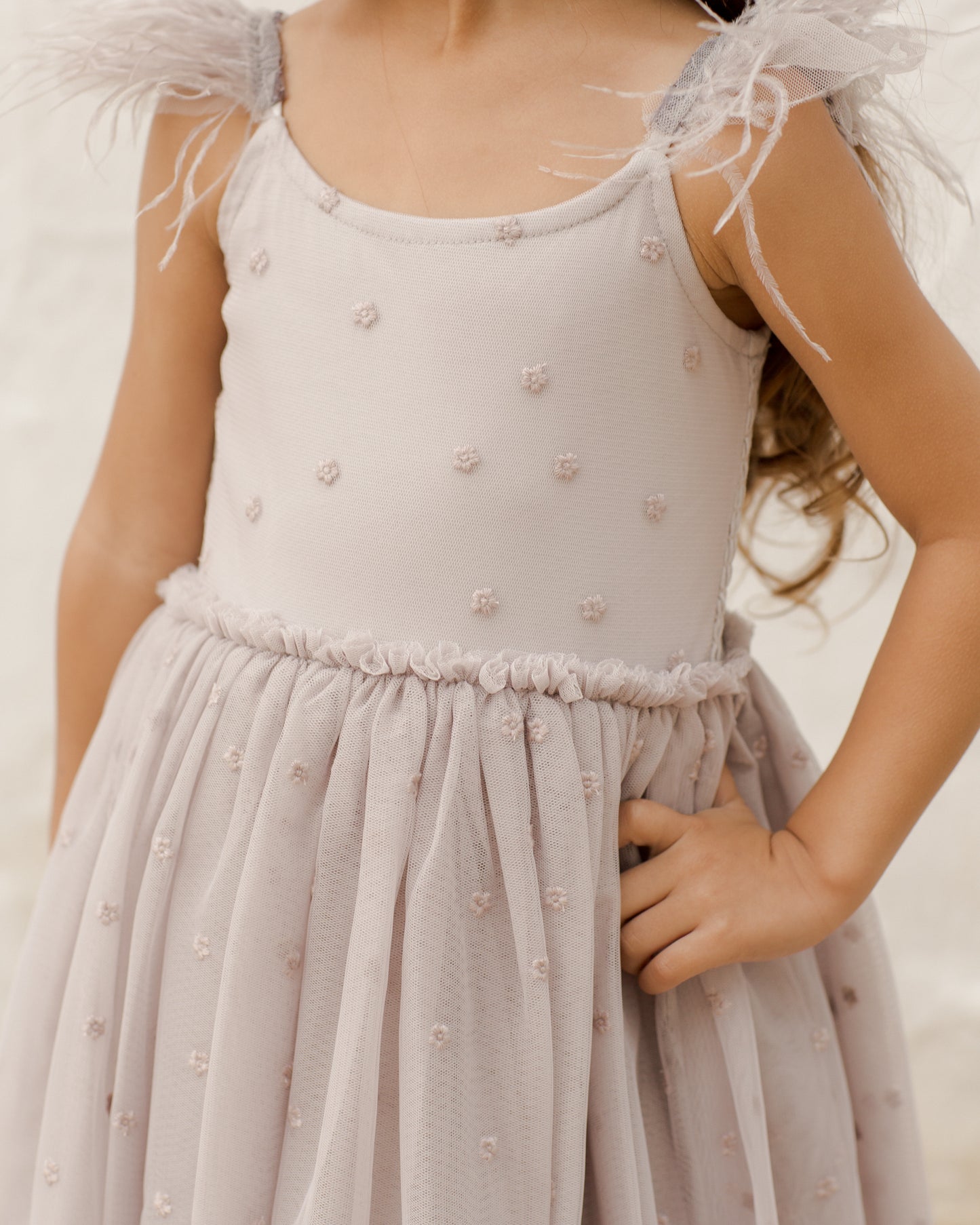 A young girl in a light pink dress with feather straps and floral embellishments, posing confidently with her hand on her hip.