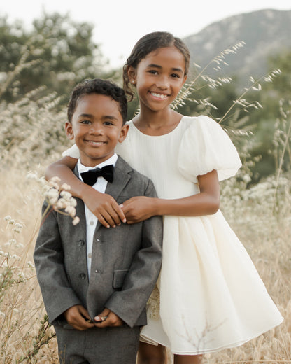 Two children pose together in a field; a boy in a suit and a girl in a white dress, smiling happily.