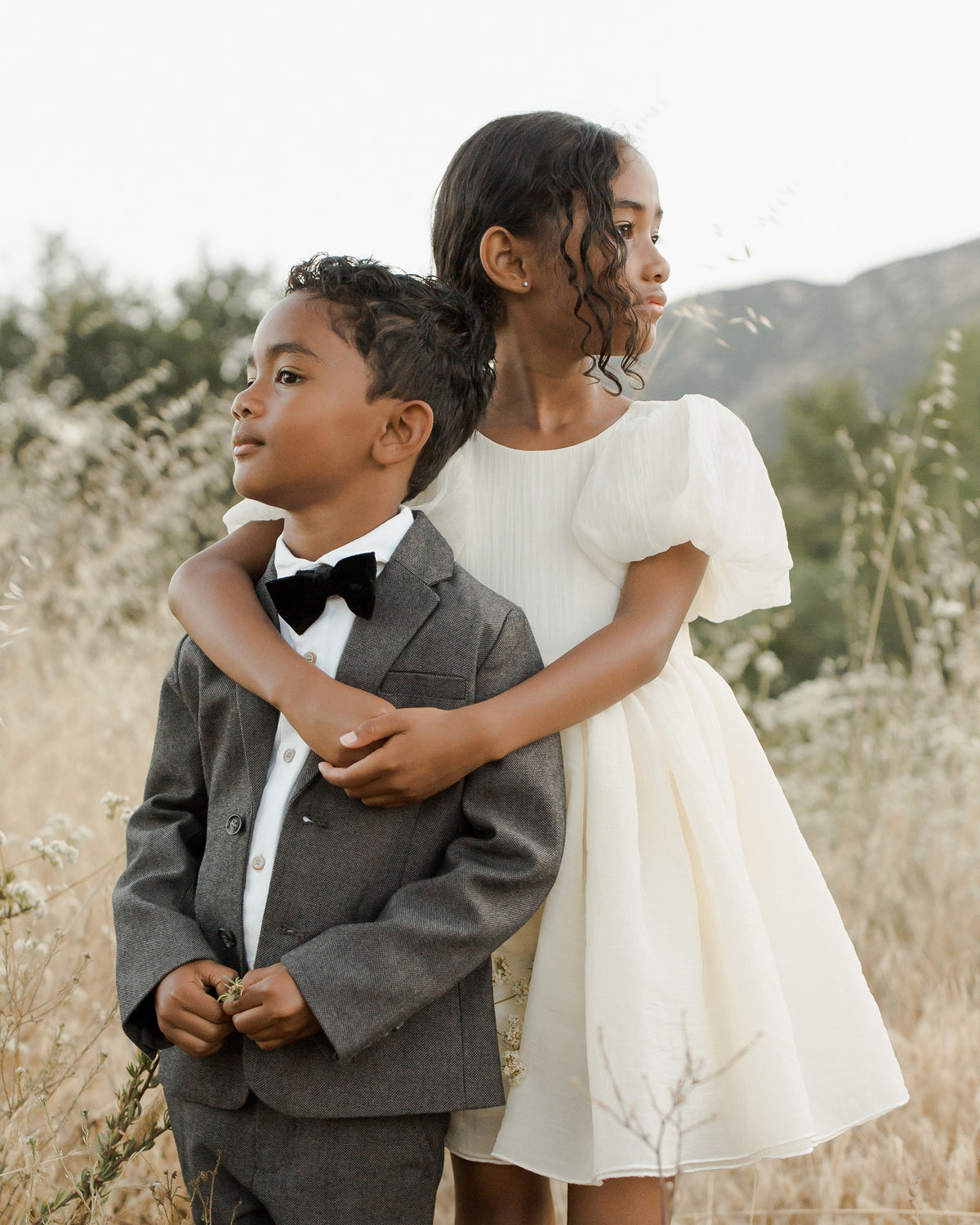 Two children stand in a field: a boy in a suit with a bow tie and a girl in a white dress, both smiling gently.