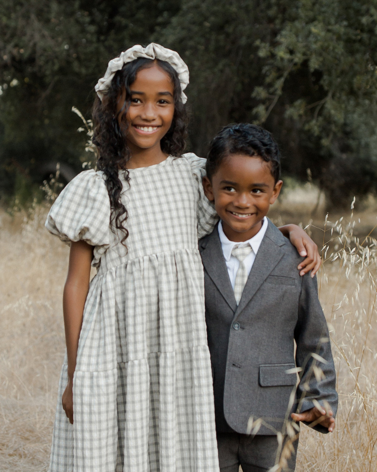 Two children stand together in a field, smiling; the girl wears a checkered dress and headband, the boy in a suit.