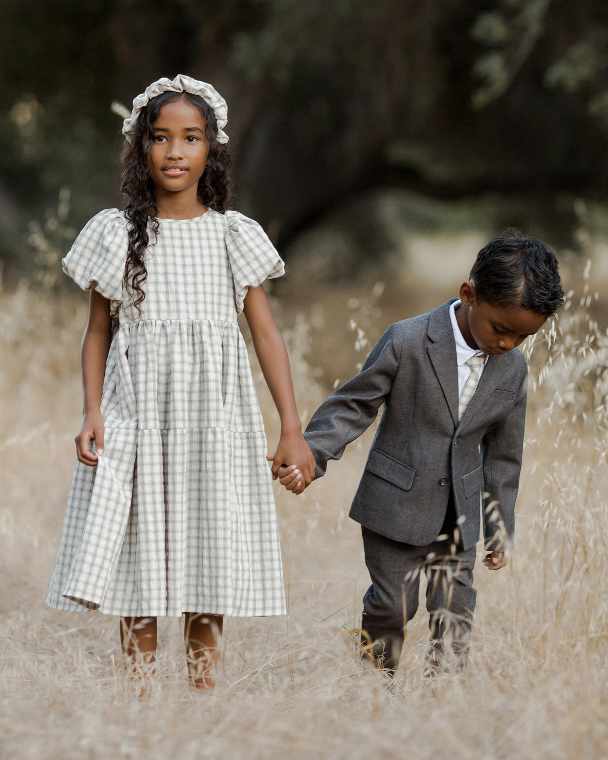 Two children hold hands in a field, wearing a dress and suit, surrounded by tall golden grass.