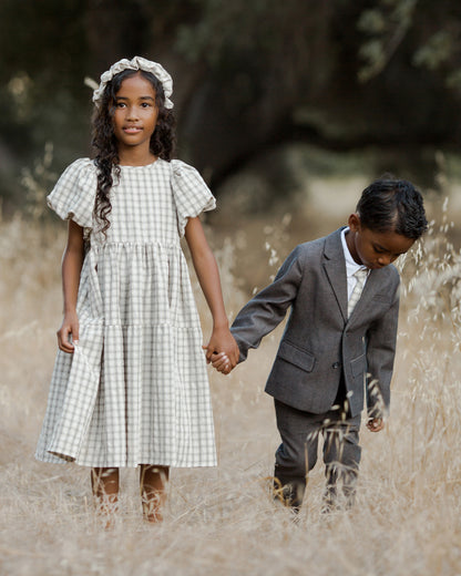 Two children hold hands in a field, wearing a dress and suit, surrounded by tall golden grass.