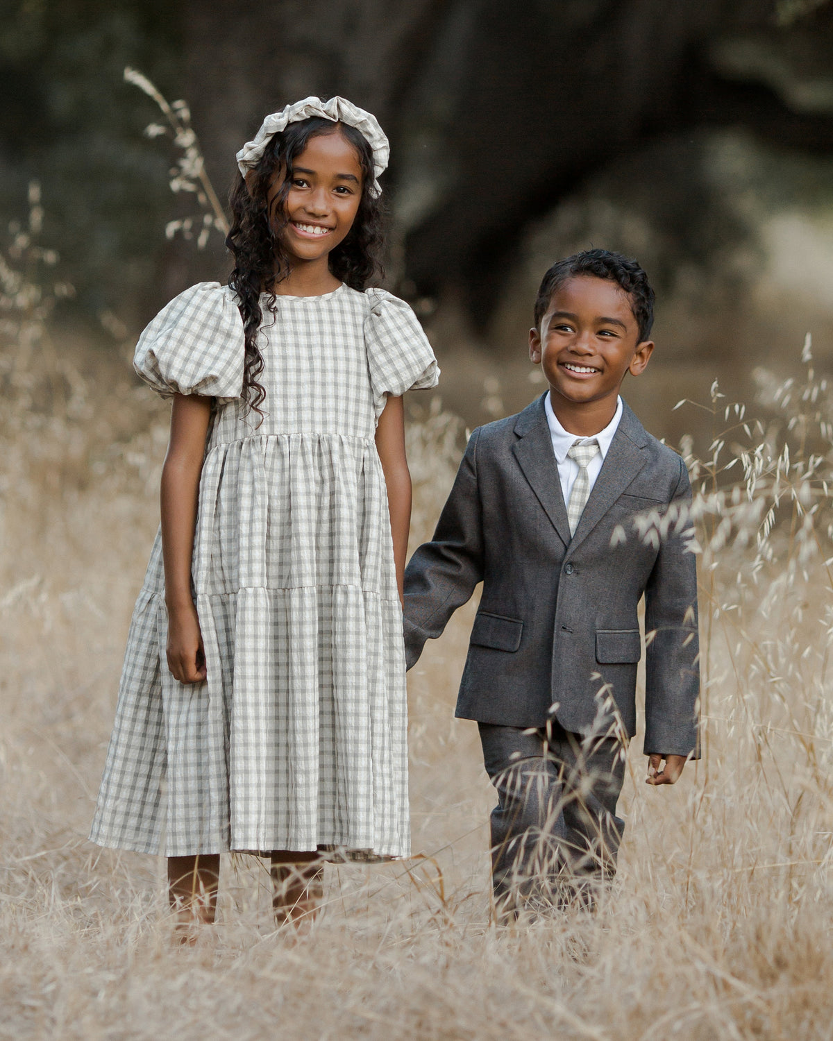 Two cheerful children stand in a grassy field, a girl in a checkered dress and a boy in a suit, smiling together.