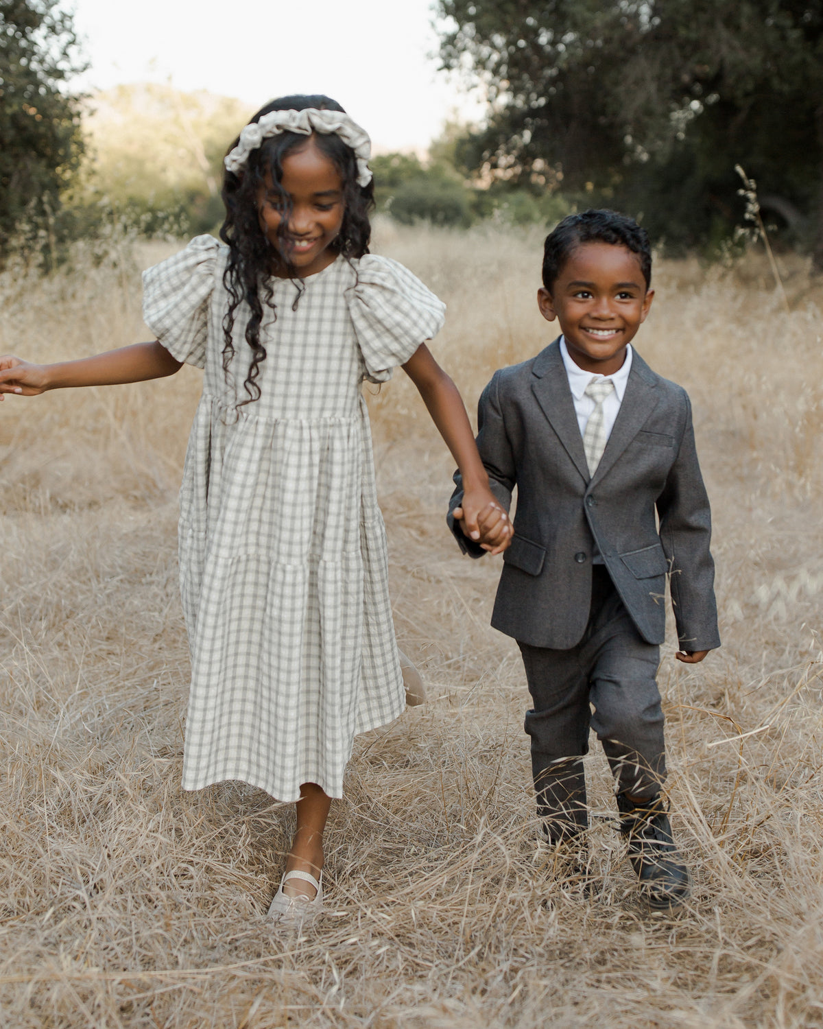 Two children walk hand in hand through a field, smiling. The girl wears a plaid dress, and the boy is in a suit.