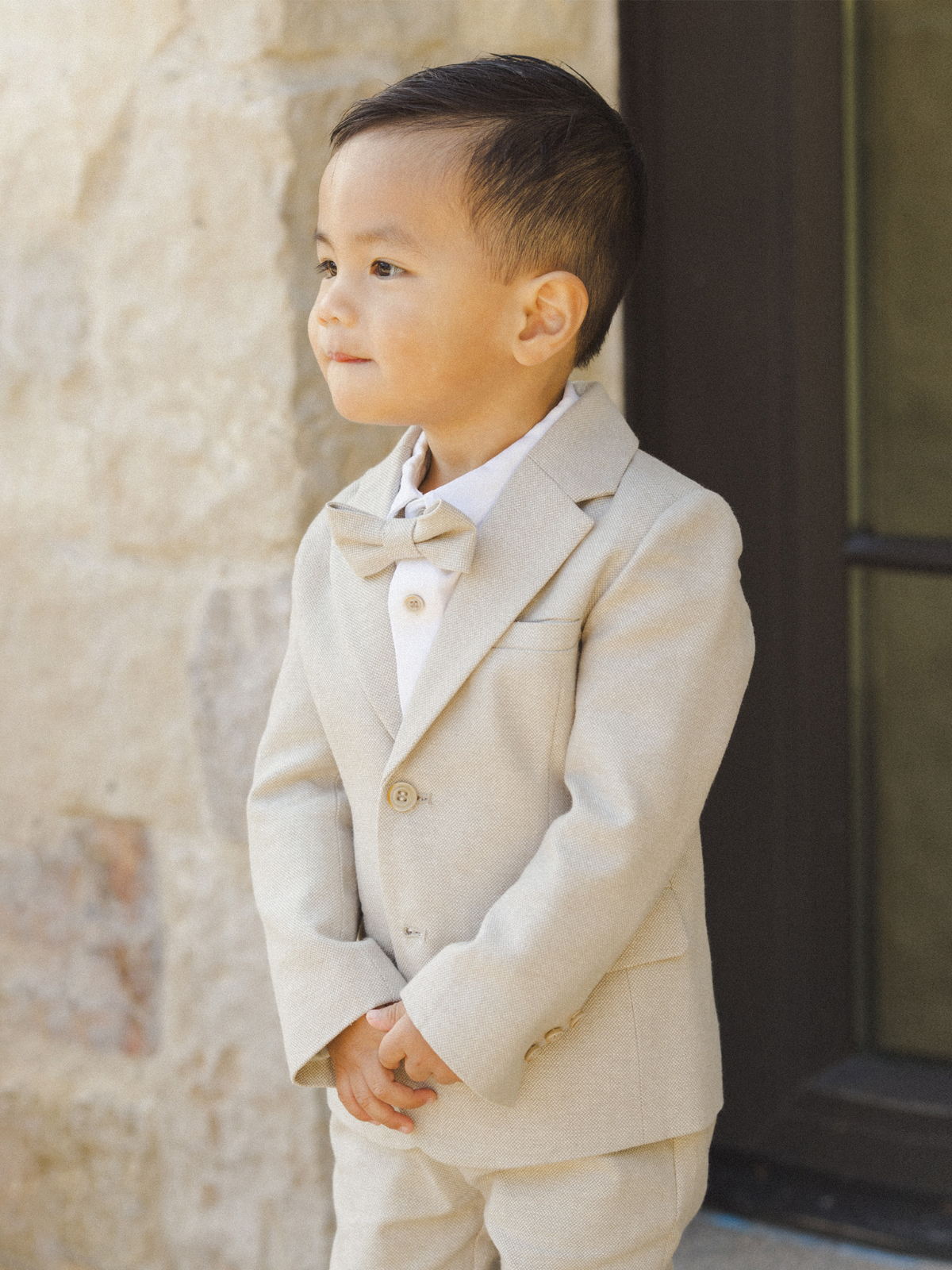 Young boy in a light gray suit and bow tie, standing with hands clasped, against a stone background.