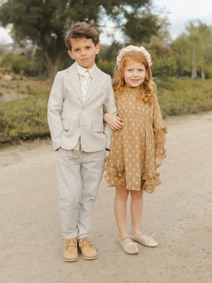 Two children, a boy in a grey suit and a girl in a floral dress, stand together on a dirt path.