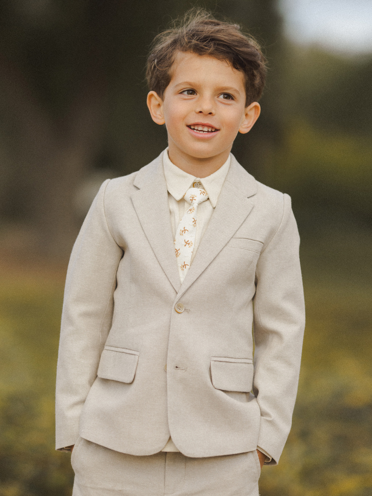 Smiling boy in a light suit with a patterned tie, standing outdoors in a natural setting.