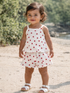 Smiling toddler in a red polka dot outfit stands on a sandy beach with greenery in the background.
