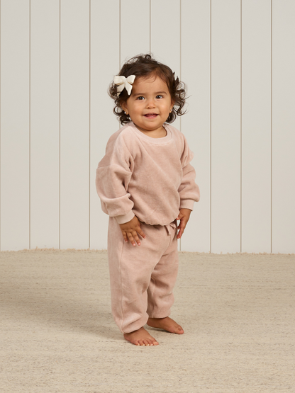 Toddler with curly hair and a bow, wearing a pink outfit, smiling in a light-filled room with wooden accents.