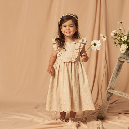 Smiling girl in cream dress holding a flower, standing on a beige backdrop next to a wooden ladder with daisies.