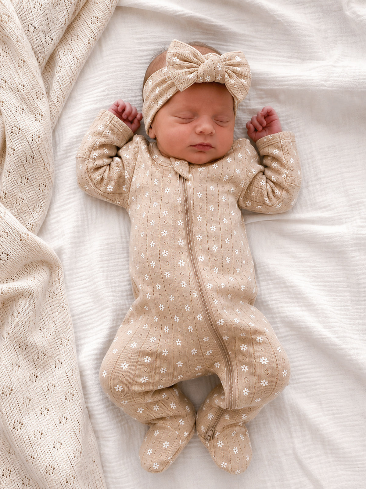 Newborn baby girl sleeping on a white blanket, wearing a beige floral onesie and matching headband.