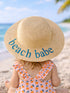 Child in a straw hat reading "beach babe," sitting on a sandy beach with ocean waves in the background.