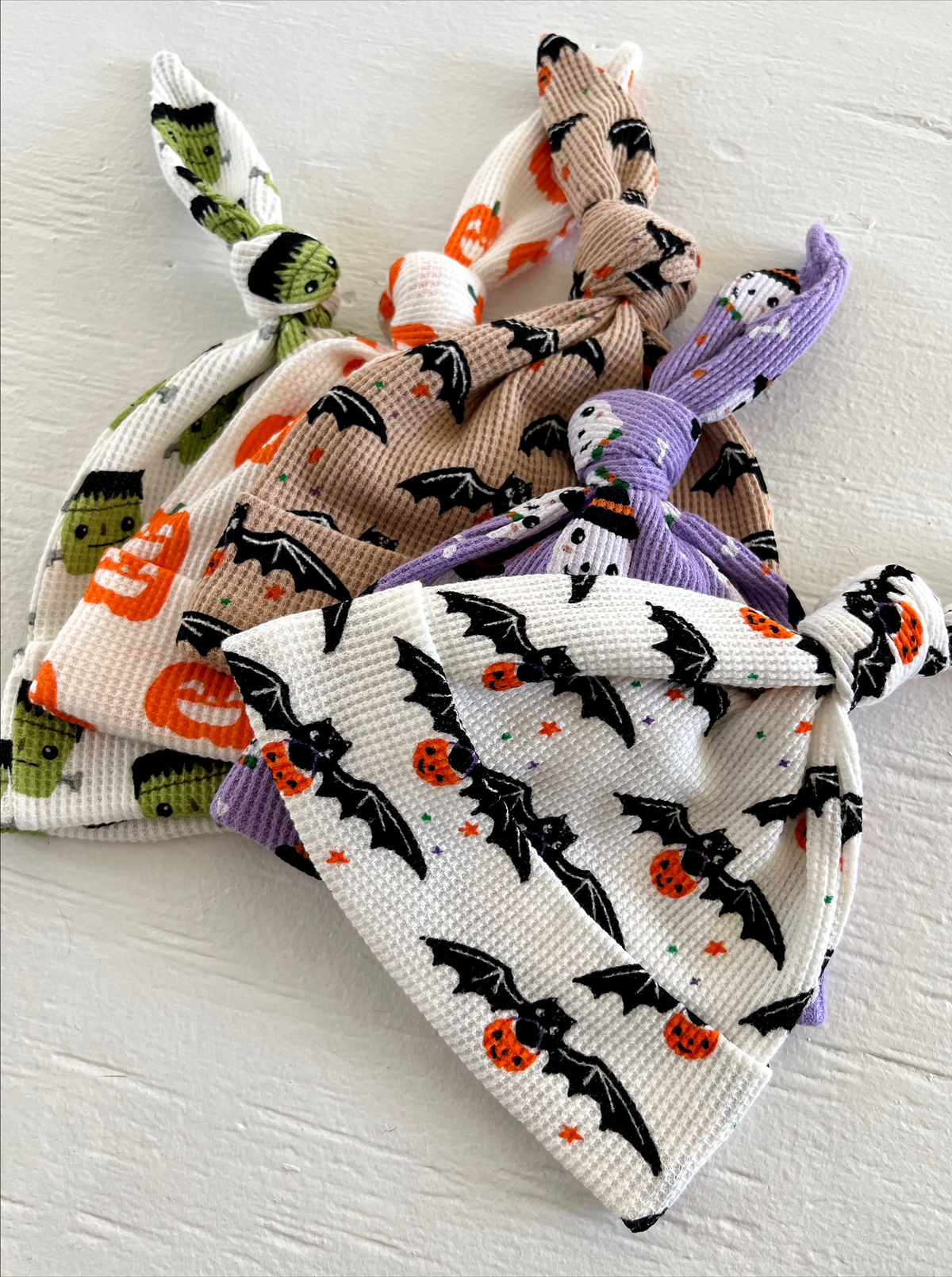 Three colorful baby hats featuring Halloween themes like bats, pumpkins, and Frankenstein, tied at the top.