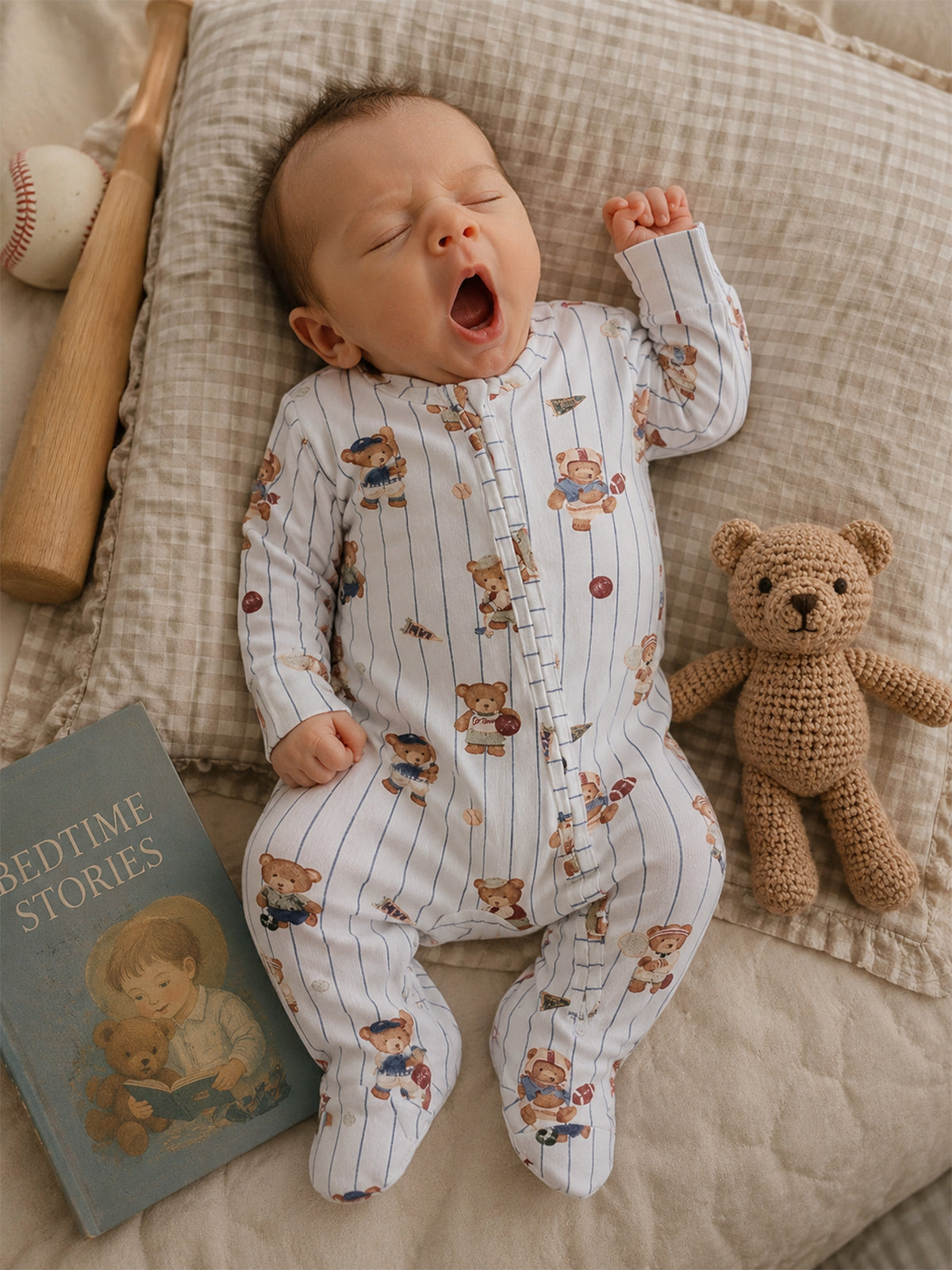 Yawning baby wearing a striped onesie with teddy bear print, surrounded by a book and a plush teddy bear.