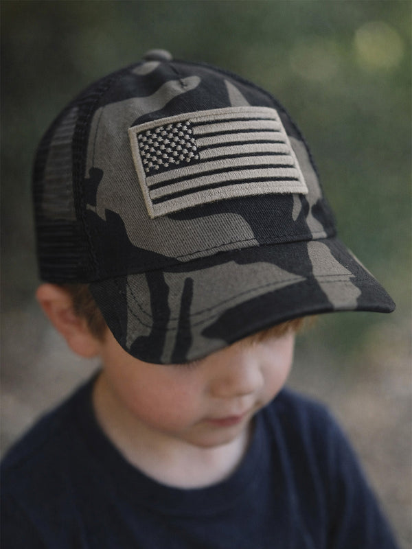 Child wearing a camo baseball cap with an American flag patch, looking down outdoors.