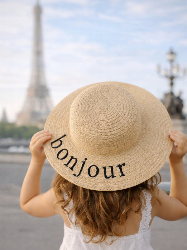 Child wearing a straw hat with "bonjour" text, facing the Eiffel Tower in a scenic outdoor setting.