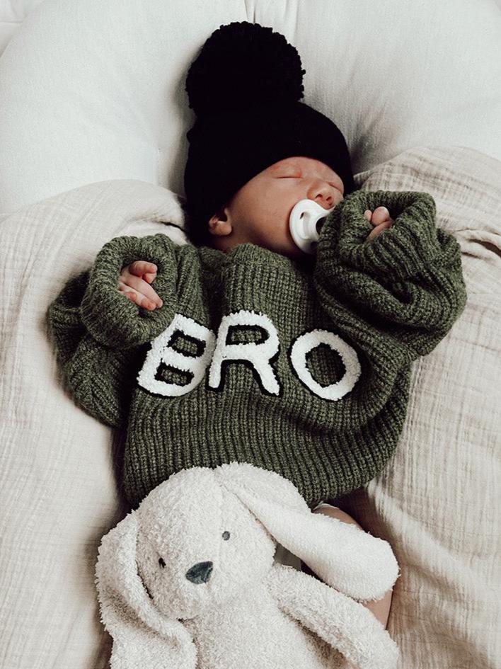 Baby wearing a green "BRO" sweater and black hat, cuddling a soft bunny toy while sleeping peacefully.