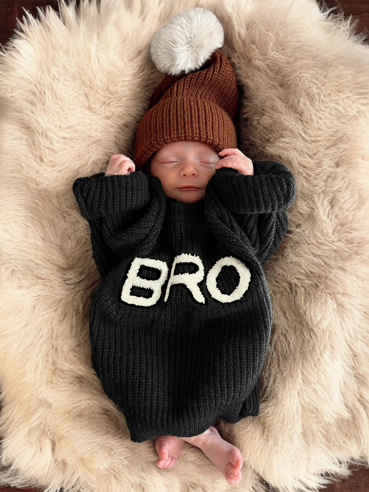 Newborn baby sleeping on a fluffy rug, wearing a black sweater with "BRO" and a brown hat with a pompom.