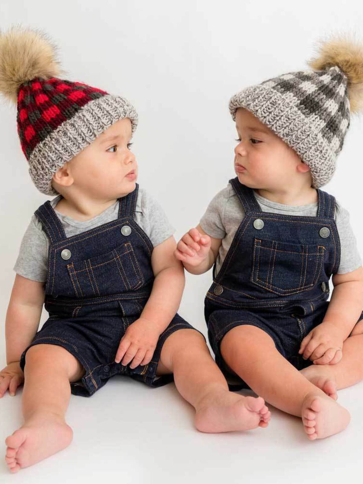 Two babies in denim overalls sit side by side, wearing knitted hats with pom-poms, sharing a moment.