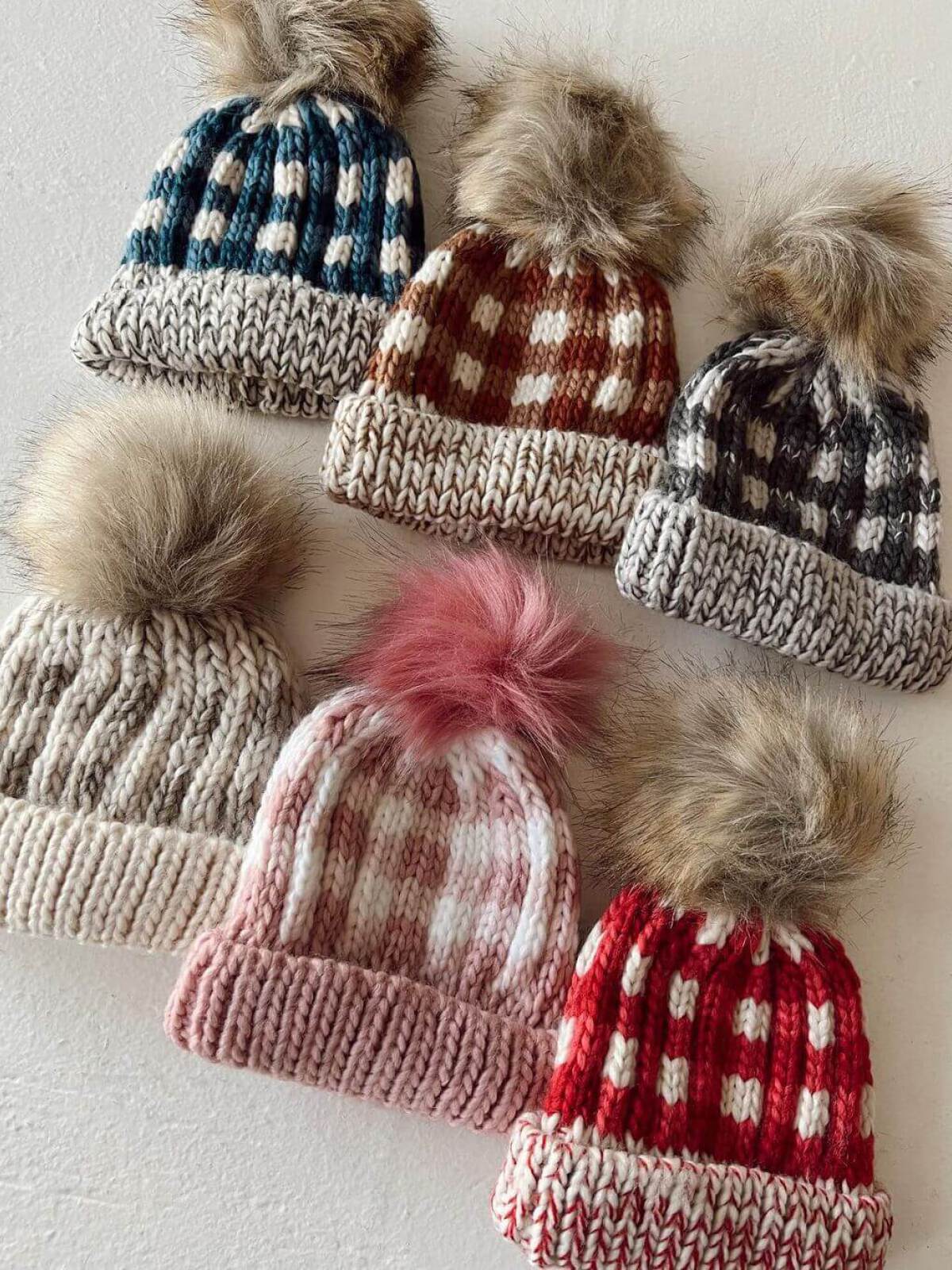 Six knitted hats with fur pom-poms in various colors and patterns laid out on a light background.