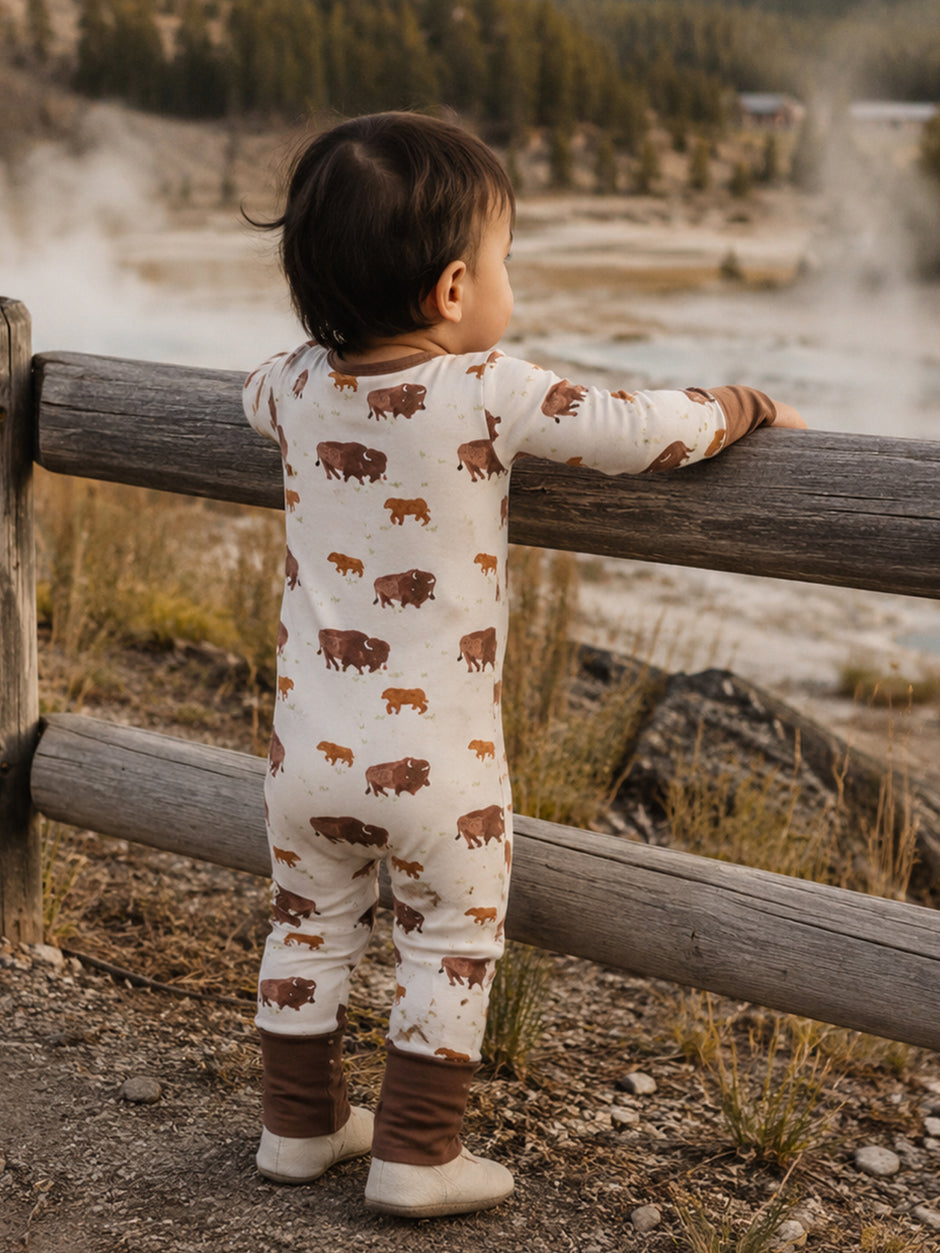 Toddler in bison-printed pajamas, leaning on a wooden fence, gazing at a natural landscape.