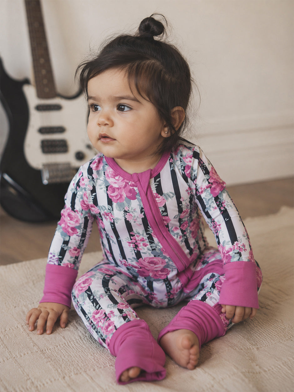 A toddler in a floral outfit sits on a blanket, looking thoughtfully to the side with a guitar in the background.
