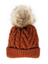 Knitted rust-orange beanie with a plush tan pom-pom on top, featuring a textured cable design.