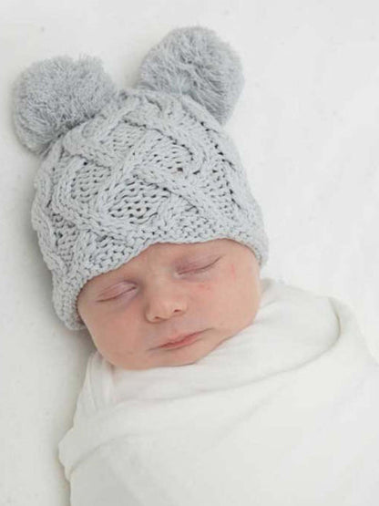 Baby sleeping peacefully, wearing a light gray knitted hat with pom-poms, wrapped in a white blanket.