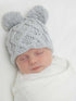 Baby sleeping peacefully, wearing a light gray knitted hat with pom-poms, wrapped in a white blanket.