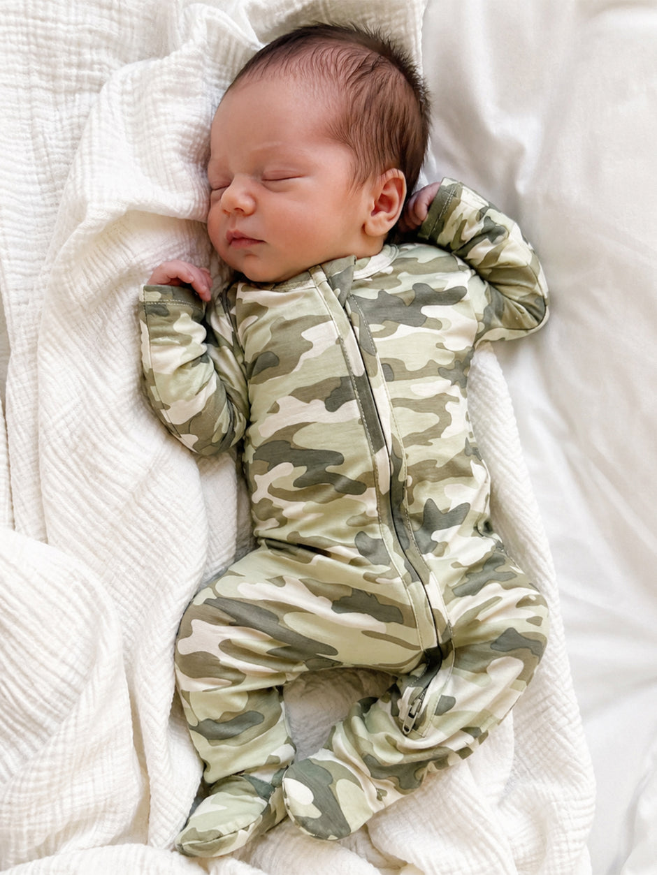 Sleepy baby in camouflage onesie resting on a soft white blanket.