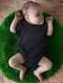 Infant sleeping peacefully on a green rug, wearing a dark onesie and a pacifier, with arms relaxed and legs spread.