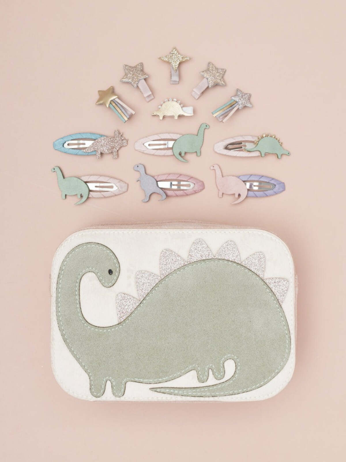 Dinosaur-themed hair clips arranged above a soft case with a dinosaur design, on a light pink background.