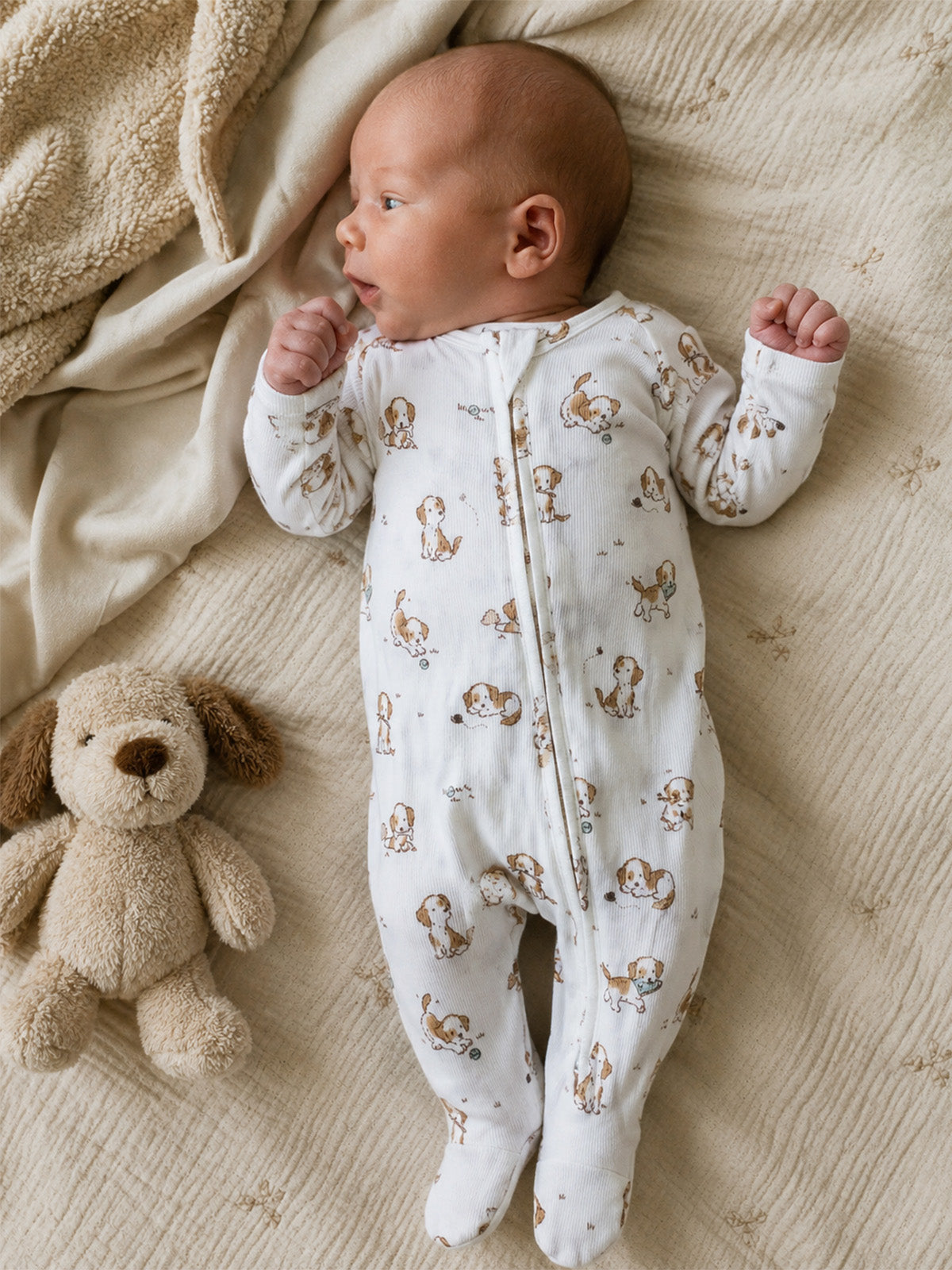 A baby in a dog-patterned onesie lies on a soft blanket next to a plush dog toy.