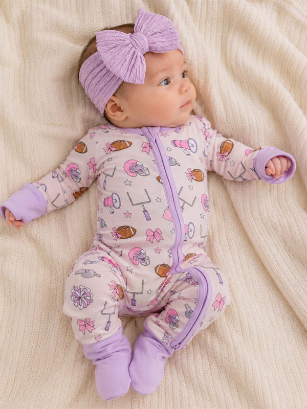 Infant girl in a purple-themed onesie and headband, lying on a soft cream blanket.