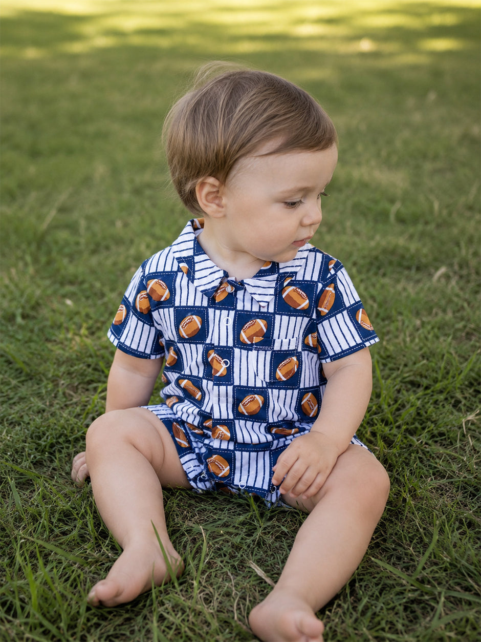 Toddler sitting on grass, wearing a navy shirt with football print and looking to the side.