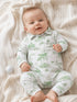 Smiling baby in green-patterned outfit on a cozy blanket, surrounded by a golf ball and soft textures.