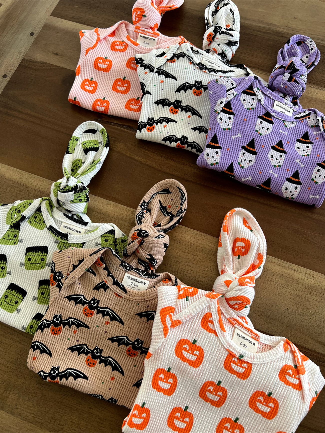 Colorful infant outfits featuring Halloween-themed designs like pumpkins, bats, and ghosts on a wooden surface.