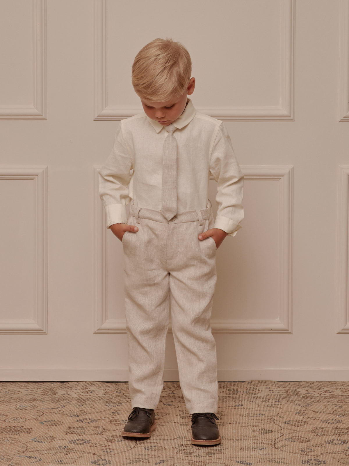 Boy in cream shirt and tie, beige trousers, and dark shoes, standing with hands in pockets against a light wall.