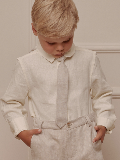 Boy in a light-colored shirt and pants, looking down, with hands in pockets against a neutral background.