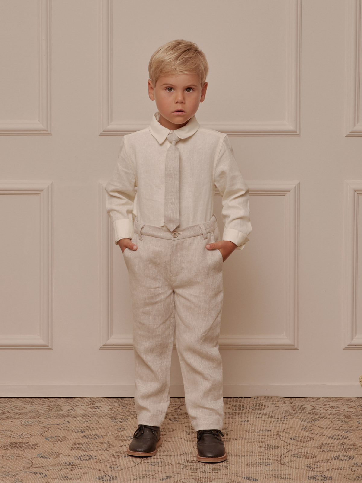 Boy wearing a cream shirt, gray tie, and beige trousers, standing confidently with hands in pockets against a neutral backdrop.