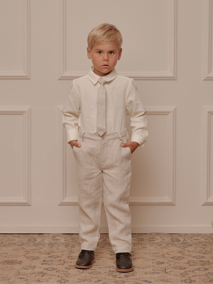 Boy wearing a cream shirt, gray tie, and beige trousers, standing confidently with hands in pockets against a neutral backdrop.