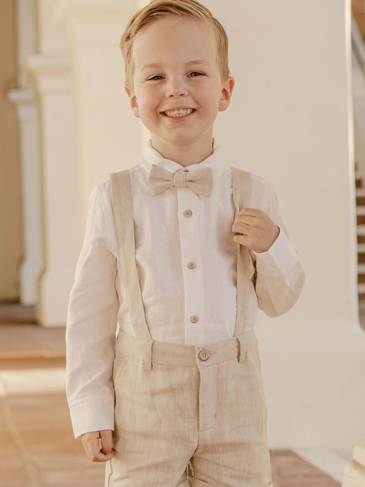 Smiling boy in a light outfit with suspenders and bow tie, standing against a neutral background.