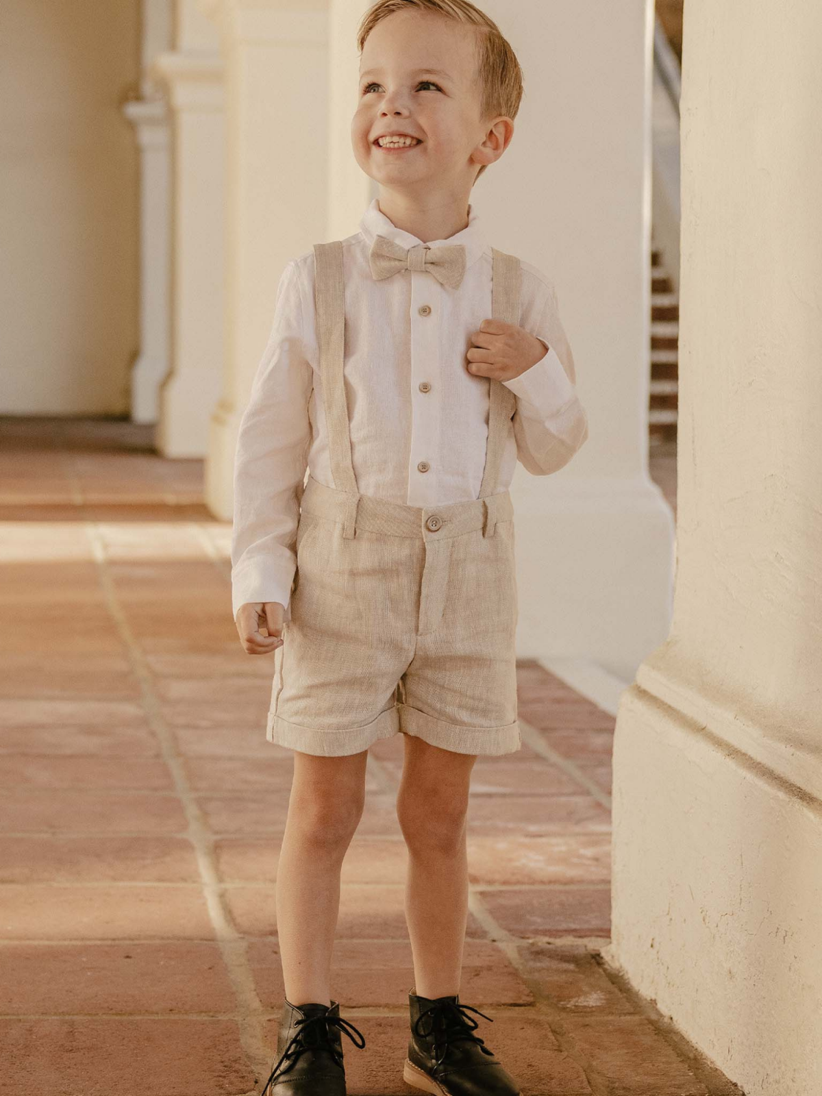 Smiling boy in a bow tie and suspenders stands in a sunny hallway, exuding playful charm.