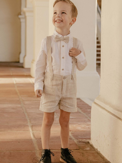Smiling boy in a bow tie and suspenders stands in a sunny hallway, exuding playful charm.