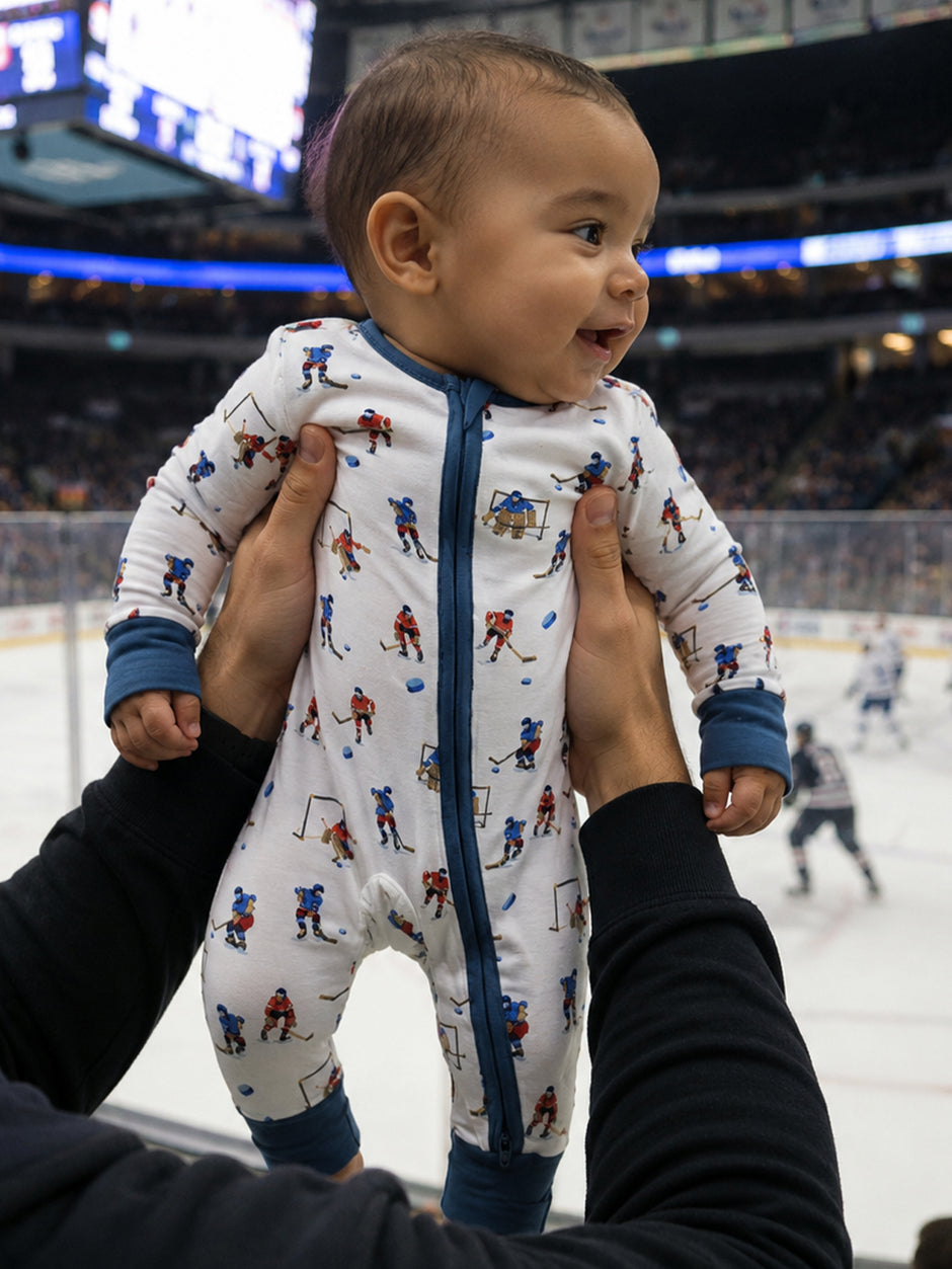 Baby in a hockey-themed onesie smiles joyfully as an adult holds them in a sports arena.