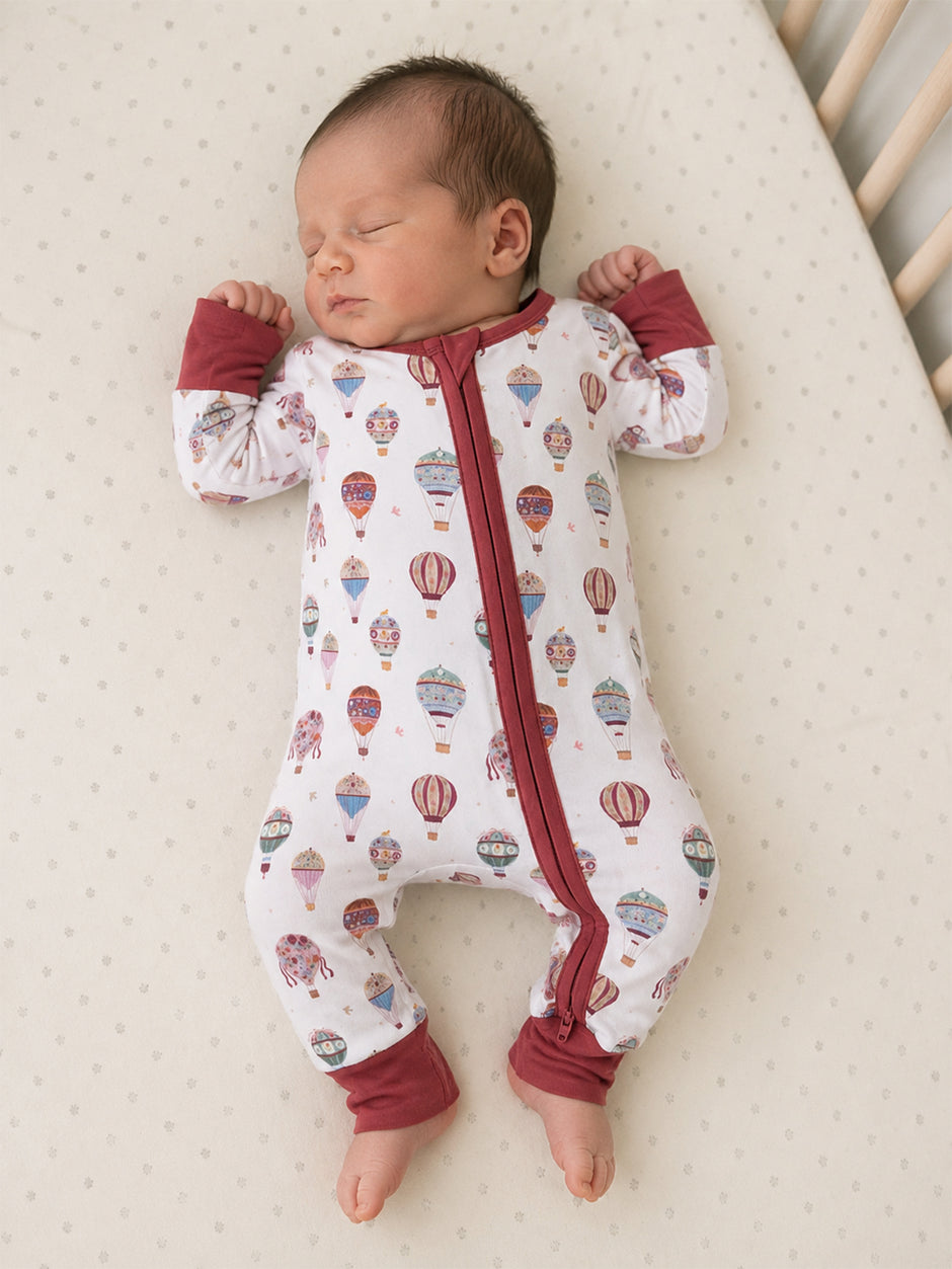 Baby sleeping in a white onesie with colorful hot air balloon prints on a light, textured mattress.